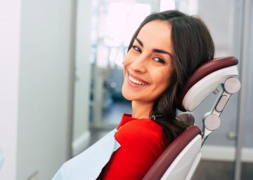 New Life With New Teeth. Gorgeous Girl Wearing Red Sweater In The Stomatology Room Full Of Day Light And White Colors Is Smiling With Her New White Eye Catching Smile.